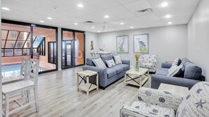 A modern living room with a grey sofa, white armchairs, and a coffee table. There is a large window with a view of a pool and a staircase leading to an upper floor. - Four Seasons 502-e Fishing Pier Newly Remodeled 3 Bedrooms (Orange Beach)