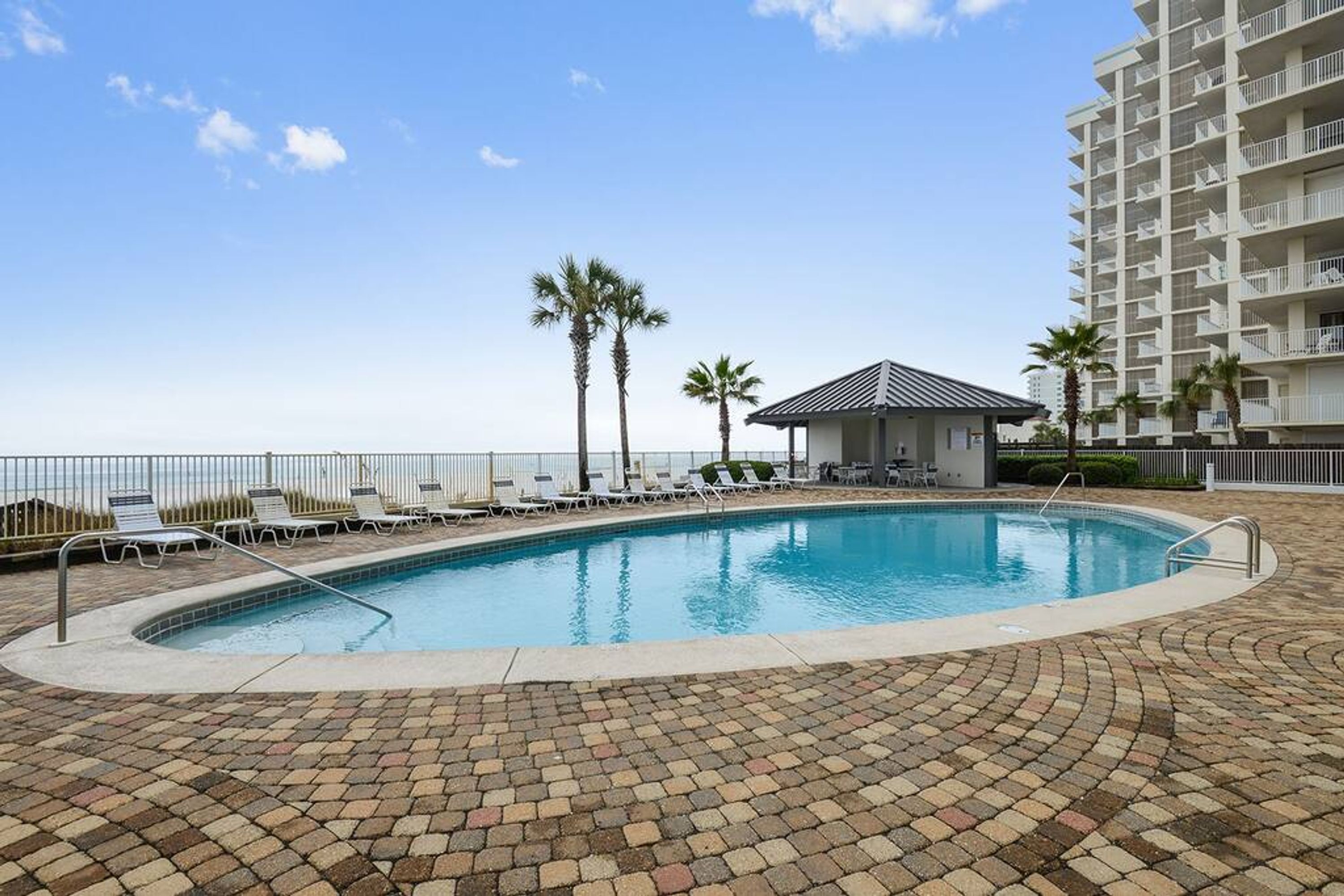 A swimming pool with lounge chairs and palm trees, surrounded by a brick patio and a high-rise building in the background.