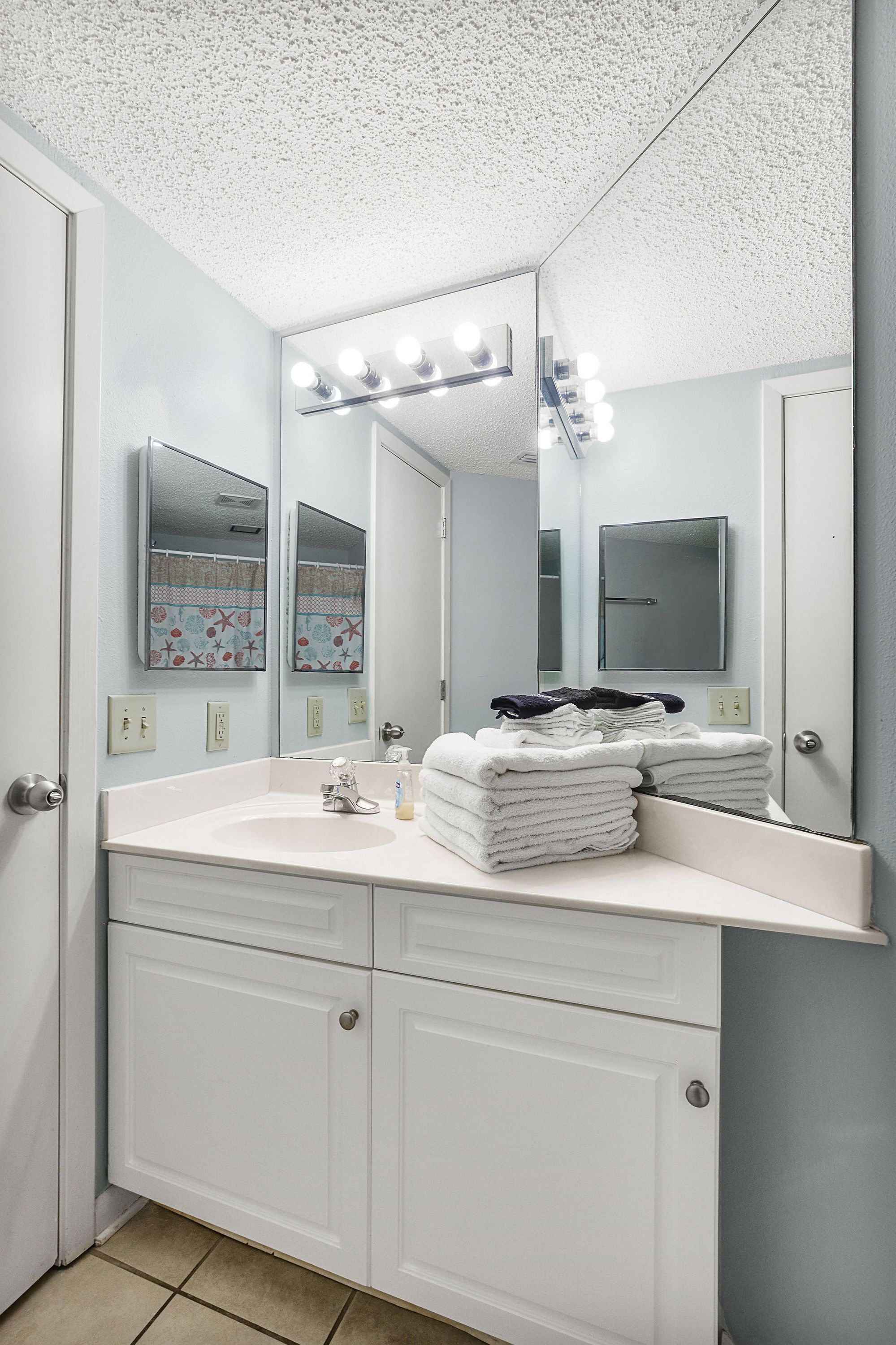 A bathroom with a white vanity, a large mirror, and a sink. There are towels stacked on the vanity, and a small shelf with toiletries.