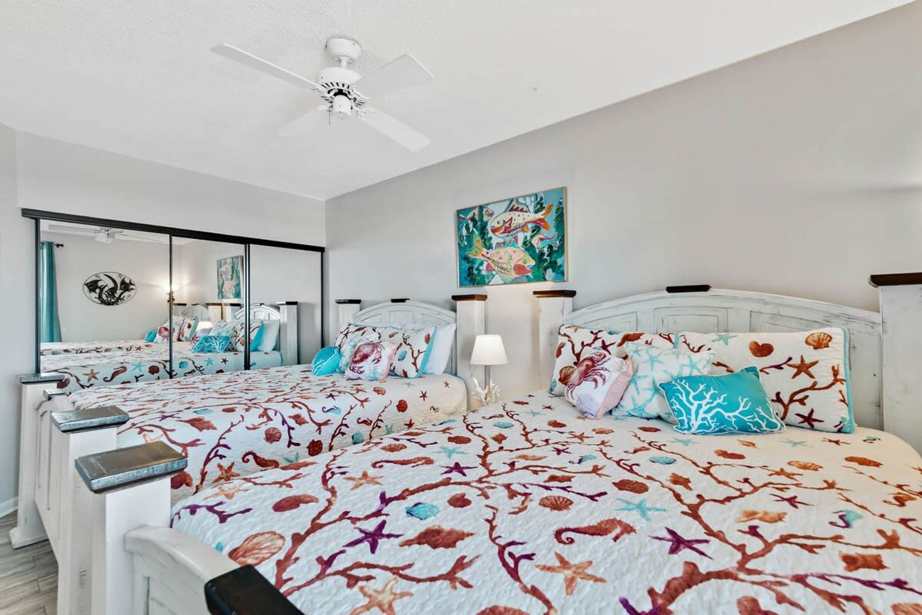 A bedroom with two beds, a ceiling fan, a large mirror, and a painting on the wall.