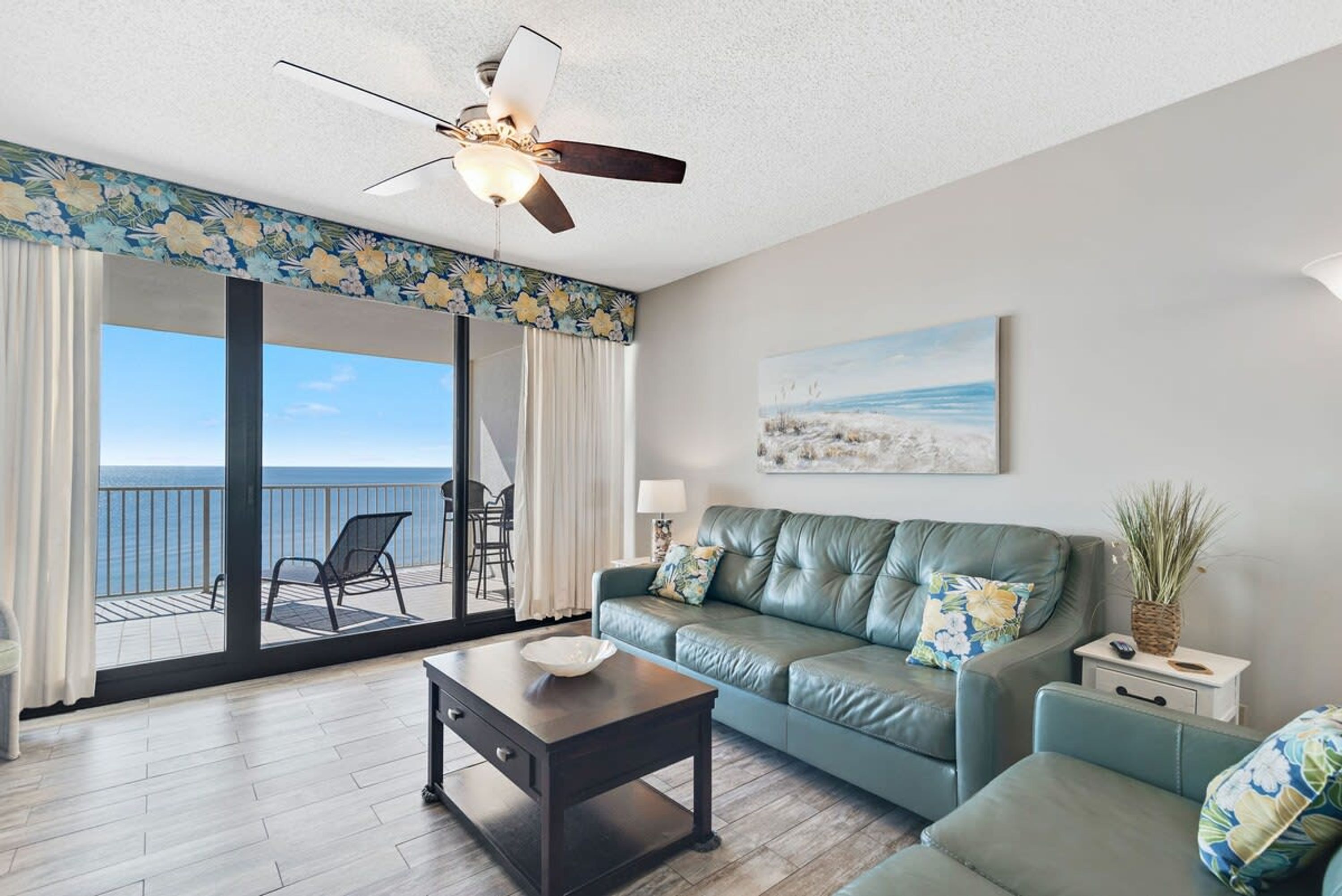 A living room with a green sofa, a coffee table, and a ceiling fan. There is a view of the ocean through large glass doors.