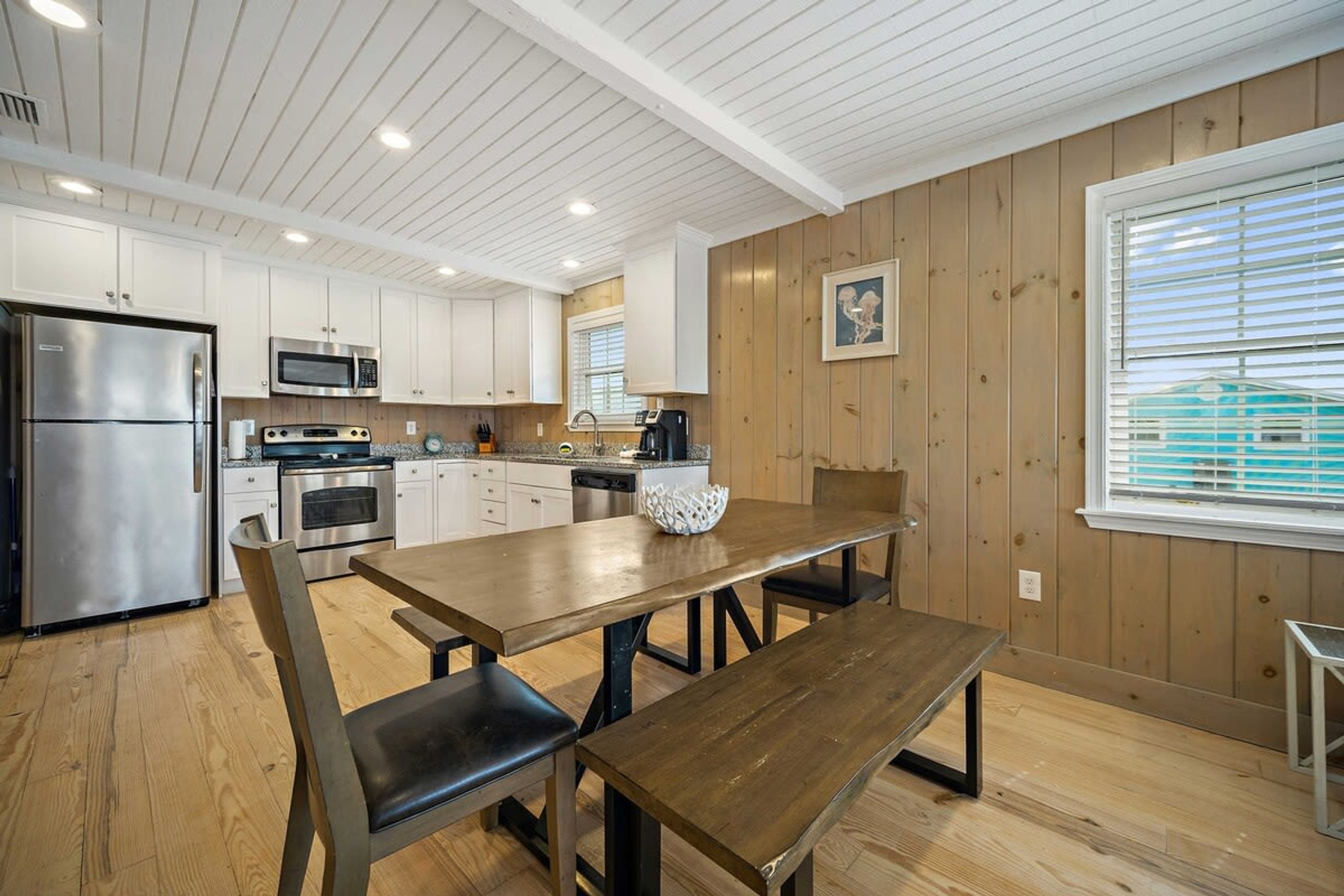 A modern kitchen with wooden walls, white cabinets, stainless steel appliances, and a wooden dining table with benches.