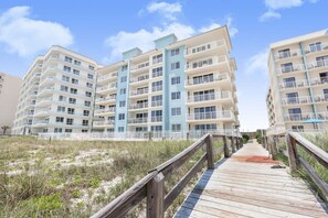 A beachfront hotel with multiple balconies, a wooden boardwalk leading to the entrance, and a clear sky. - Emerald Key 603 Beachfront Balcony 6th Floor Corner Unit (Orange Beach)