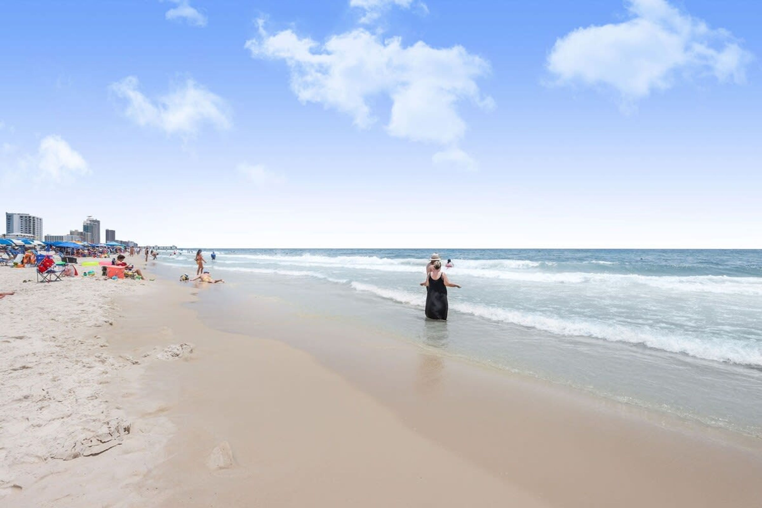 Una playa con cielo despejado, gente caminando por la orilla y sombrillas colocadas sobre la arena.