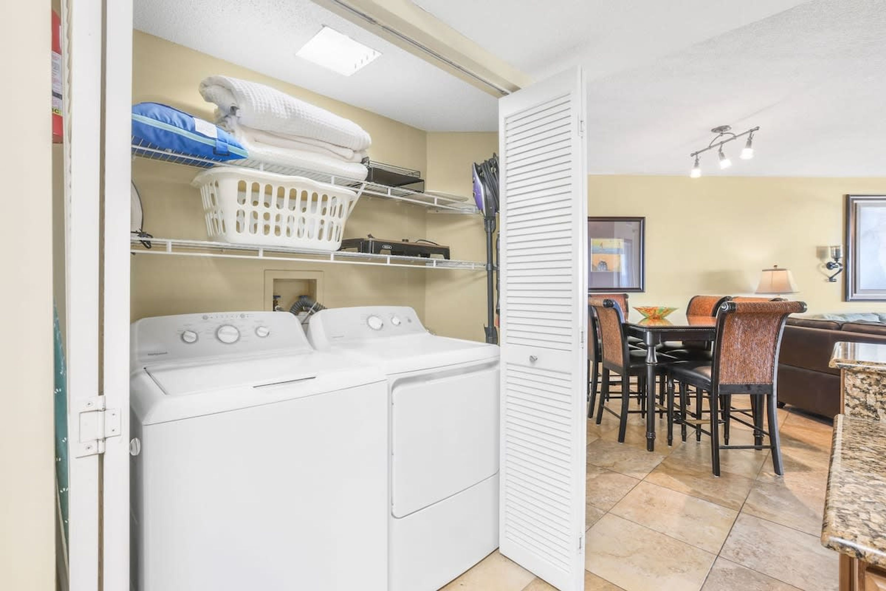 A laundry room with a washing machine, shelves with folded towels, and a dining area with chairs and a table.