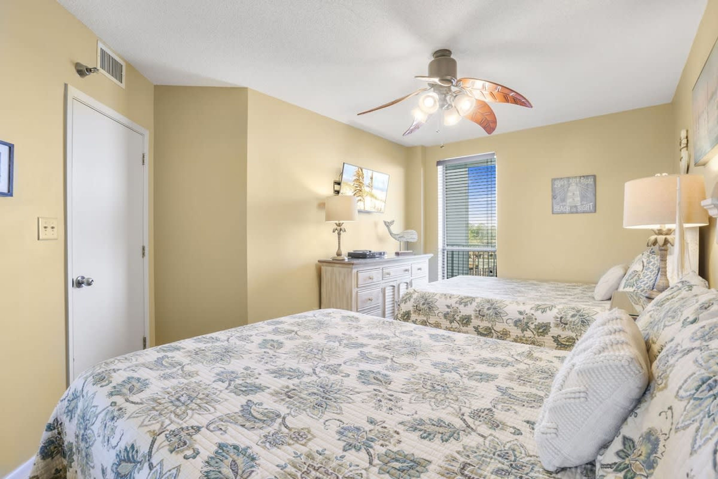 A bedroom with two beds, a ceiling fan, a dresser, and a window with blinds.