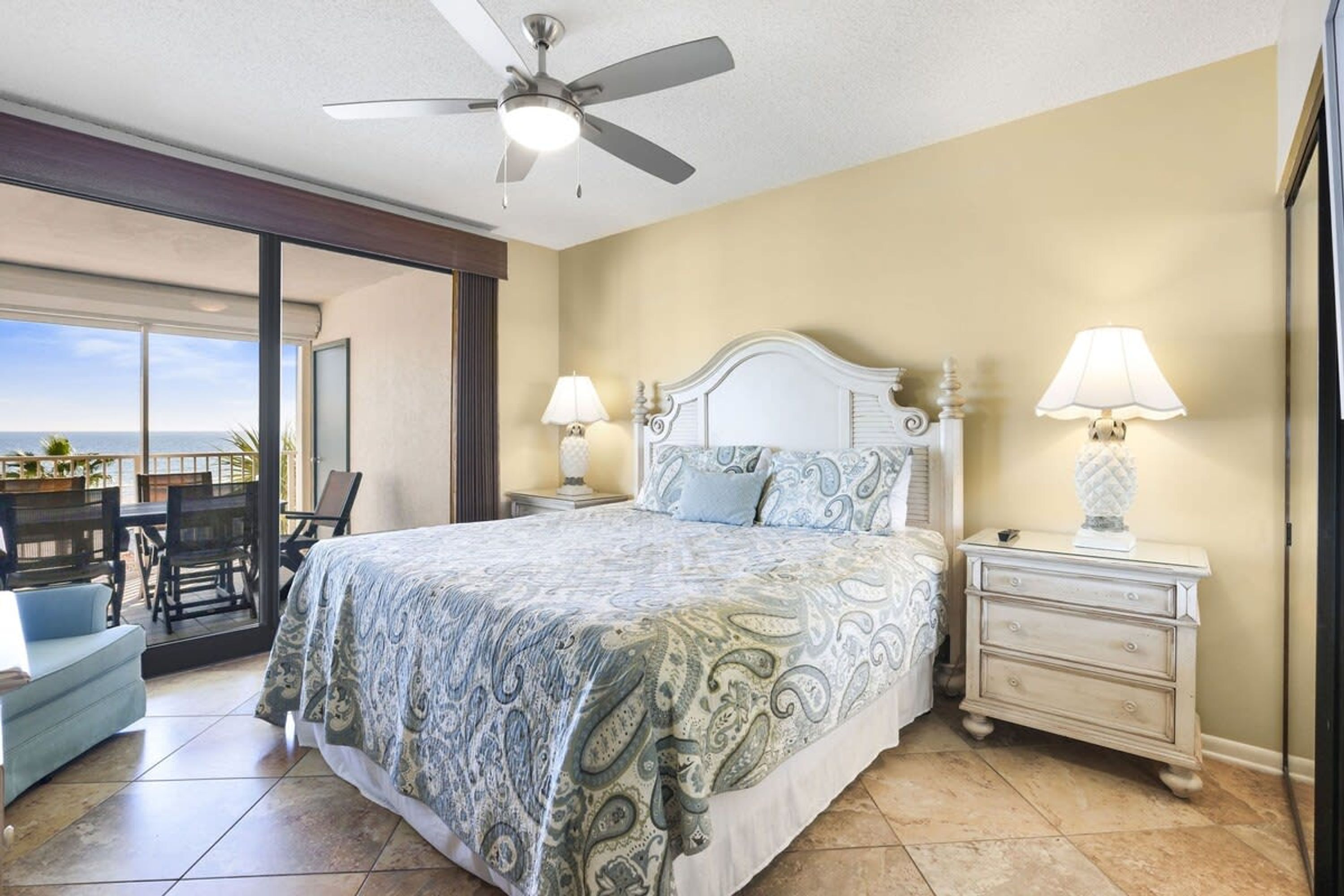 A bedroom with a large bed, two bedside tables with lamps, a ceiling fan, and a view of the ocean through a sliding door.