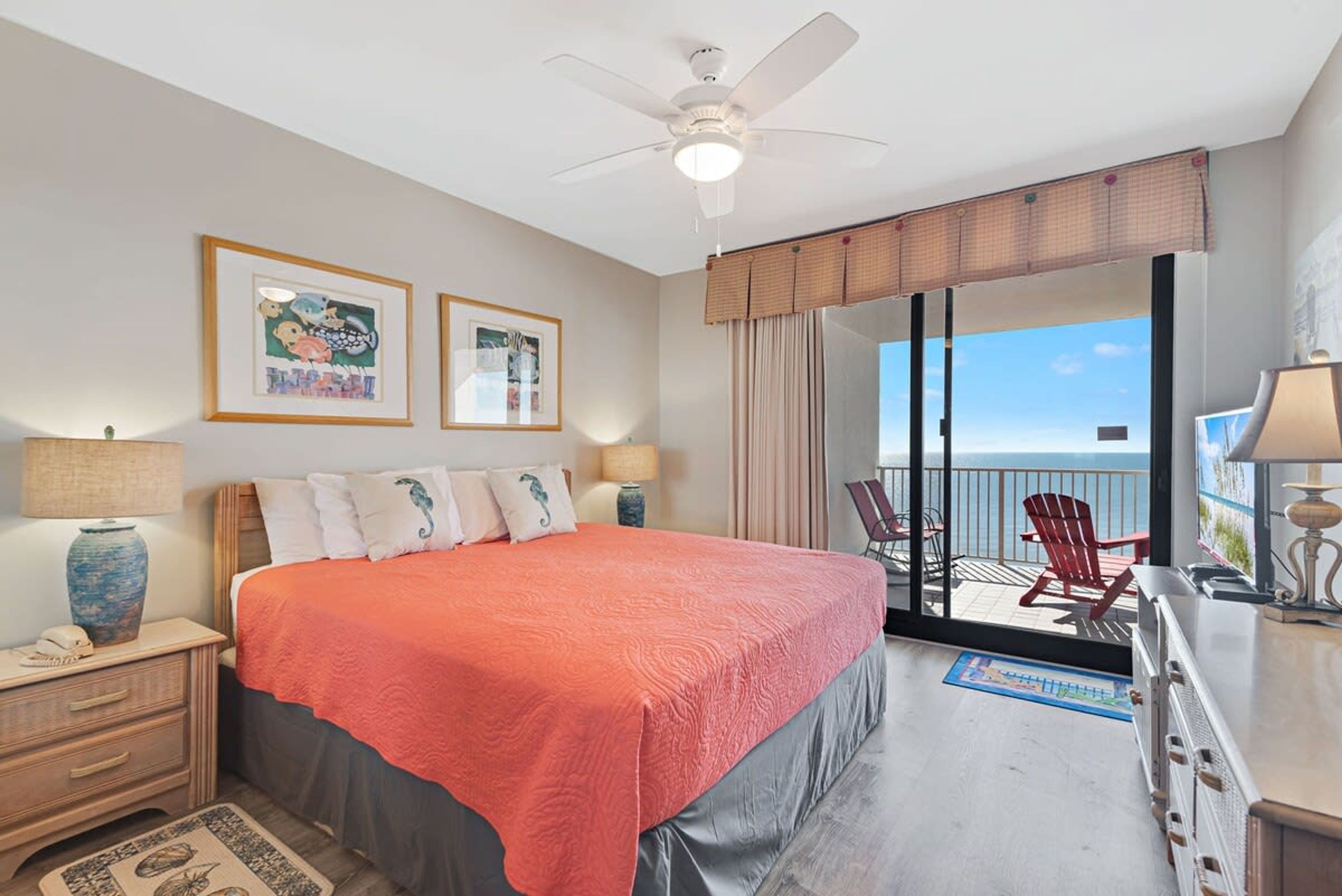 A bedroom with a large bed, a ceiling fan, a nightstand, a dresser, and a view of the balcony with a chair.