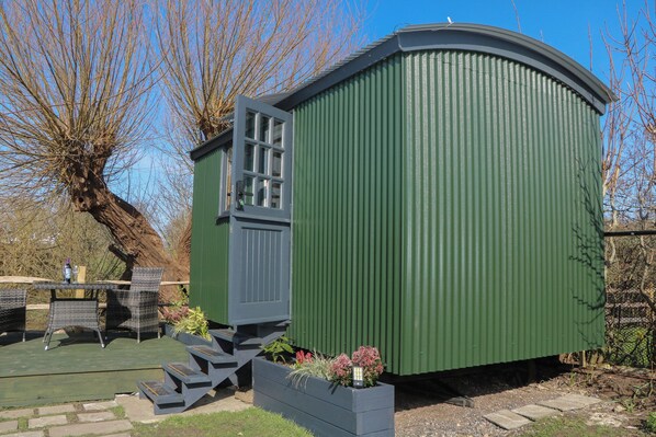 A green metal shed with a door and windows, surrounded by a garden with steps leading up to it. - Salthaven Shepherds Hut (Pevensey)