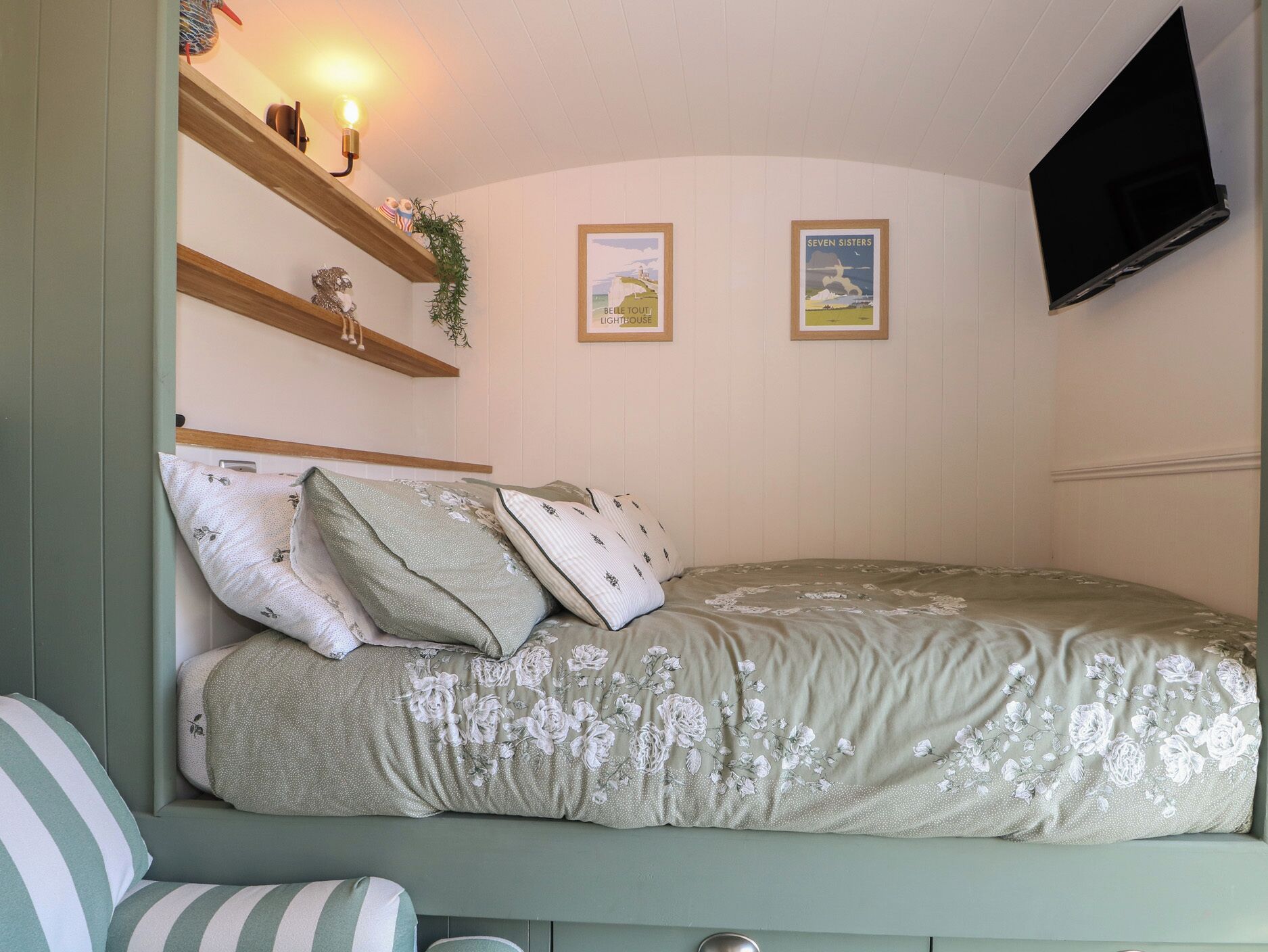 A bedroom with a bed, pillows, a shelf with decorative items, a wall-mounted TV, and framed pictures.