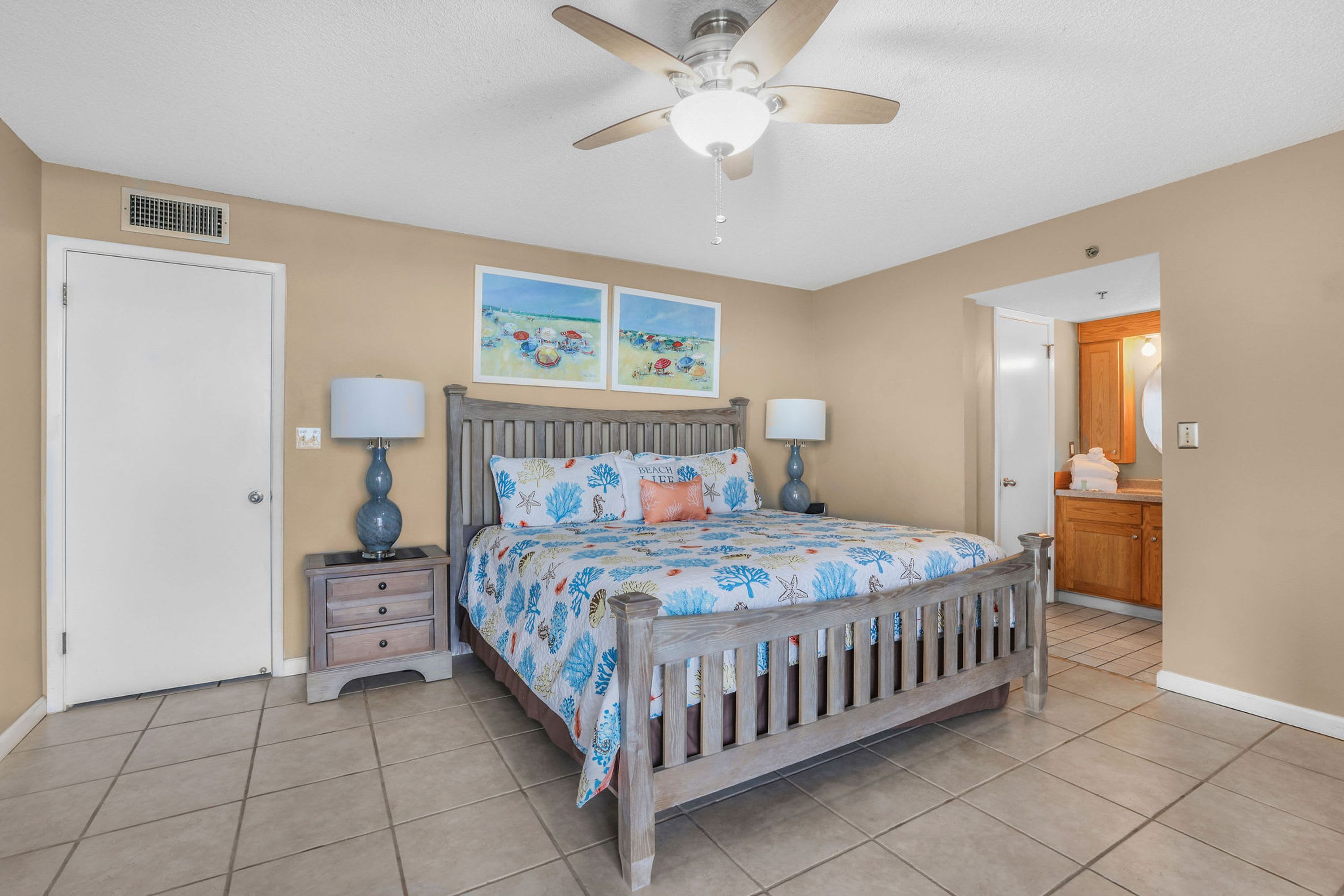 A bedroom with a wooden bed, two bedside tables with lamps, a ceiling fan, and two beach-themed wall art pieces.