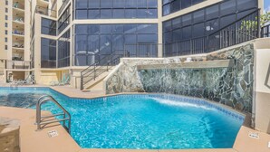A swimming pool with a stone wall and a waterfall feature, surrounded by a multi-story building with balconies. - Phoenix II 2101 3 Bedrooms Amenities Galore Beachfront (Orange Beach)