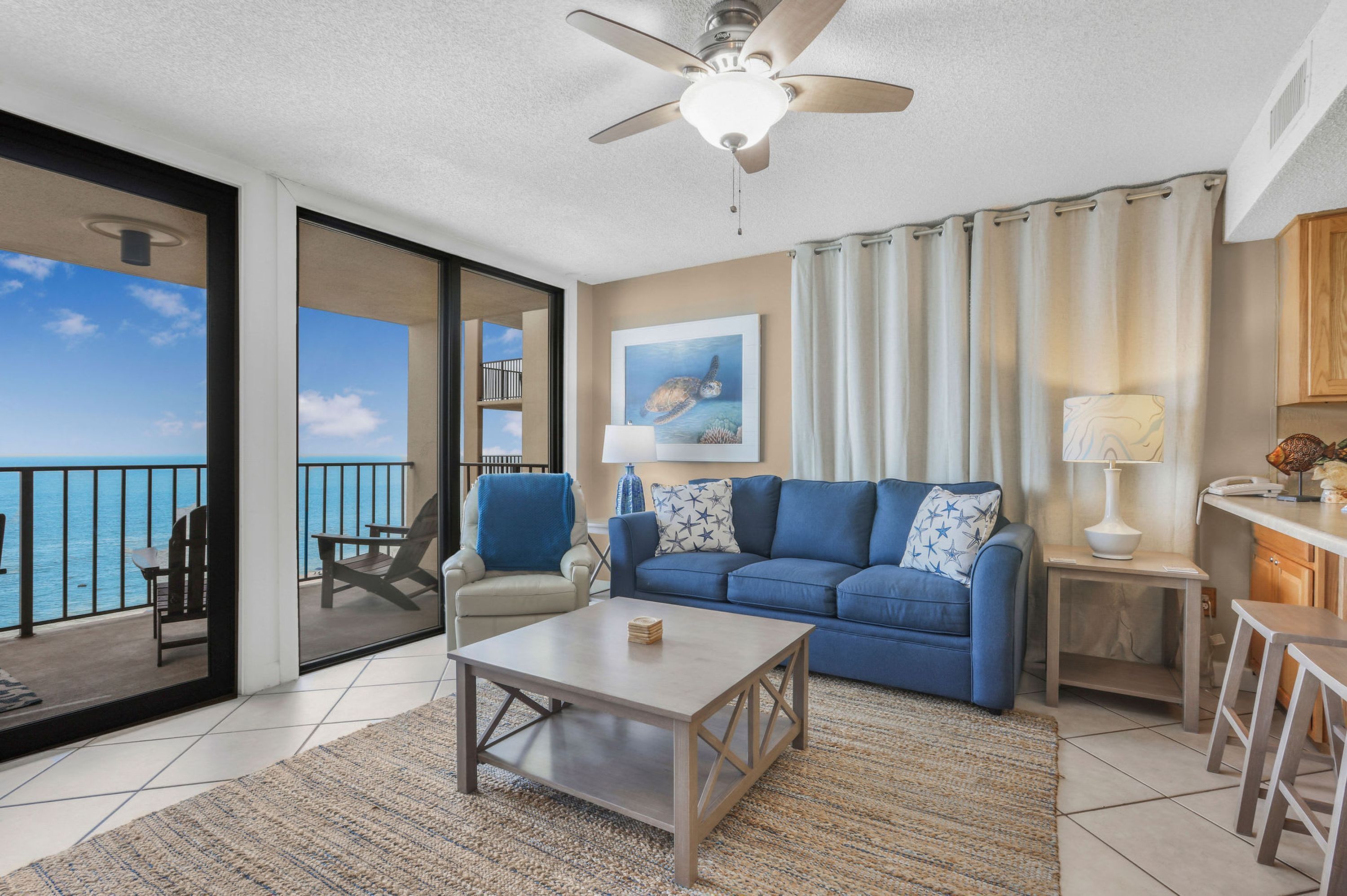 Ein Wohnzimmer mit einem blauen Sofa, einem Couchtisch, einem Deckenventilator und Blick auf das Meer durch eine Schiebetür.