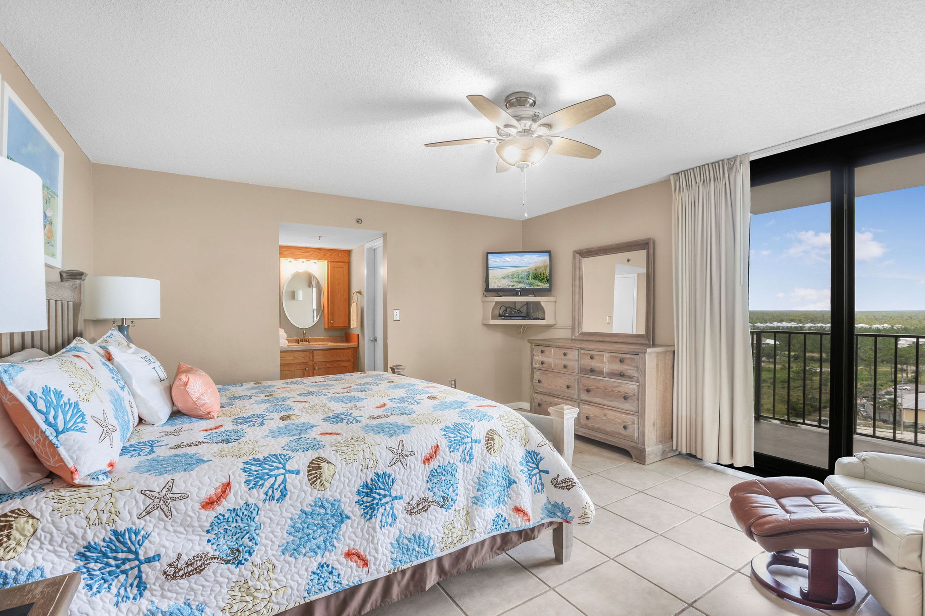 A bedroom with a bed, a ceiling fan, a dresser, a chair, and a balcony with a view.