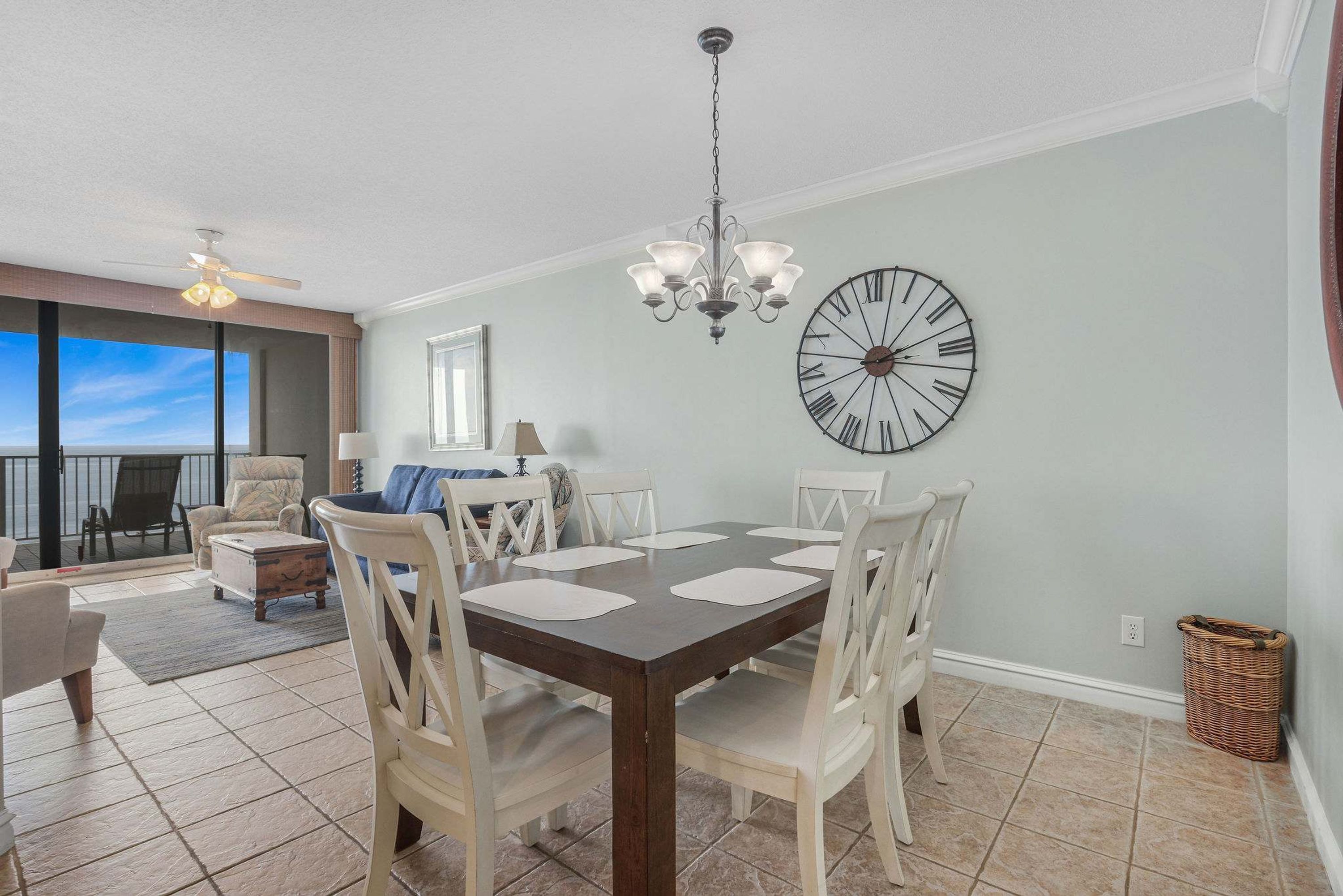 A dining area with a dark wooden table, white chairs, a chandelier, and a large wall clock.