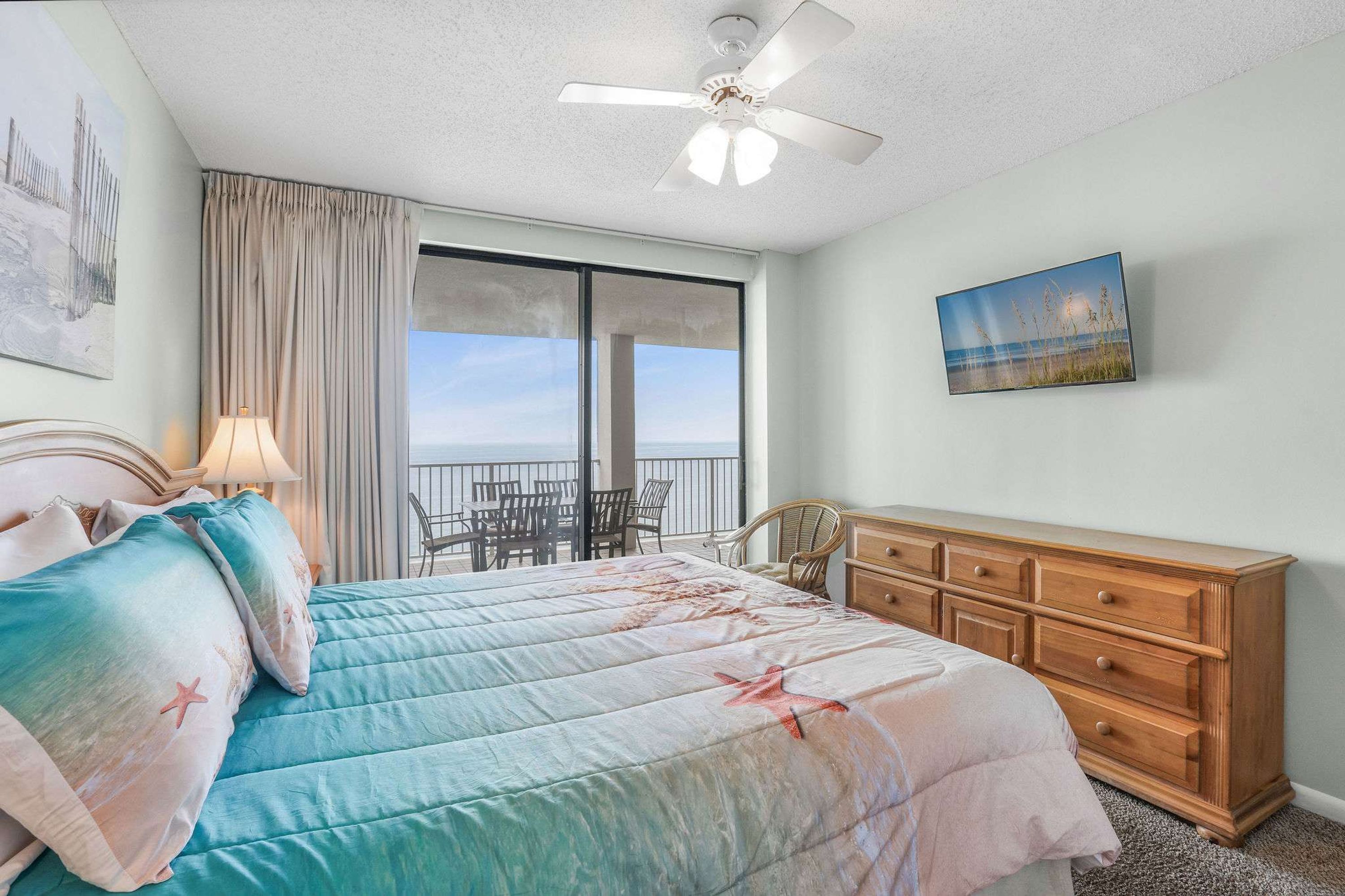 A bedroom with a bed, a dresser, a ceiling fan, a painting, and a view of the ocean.