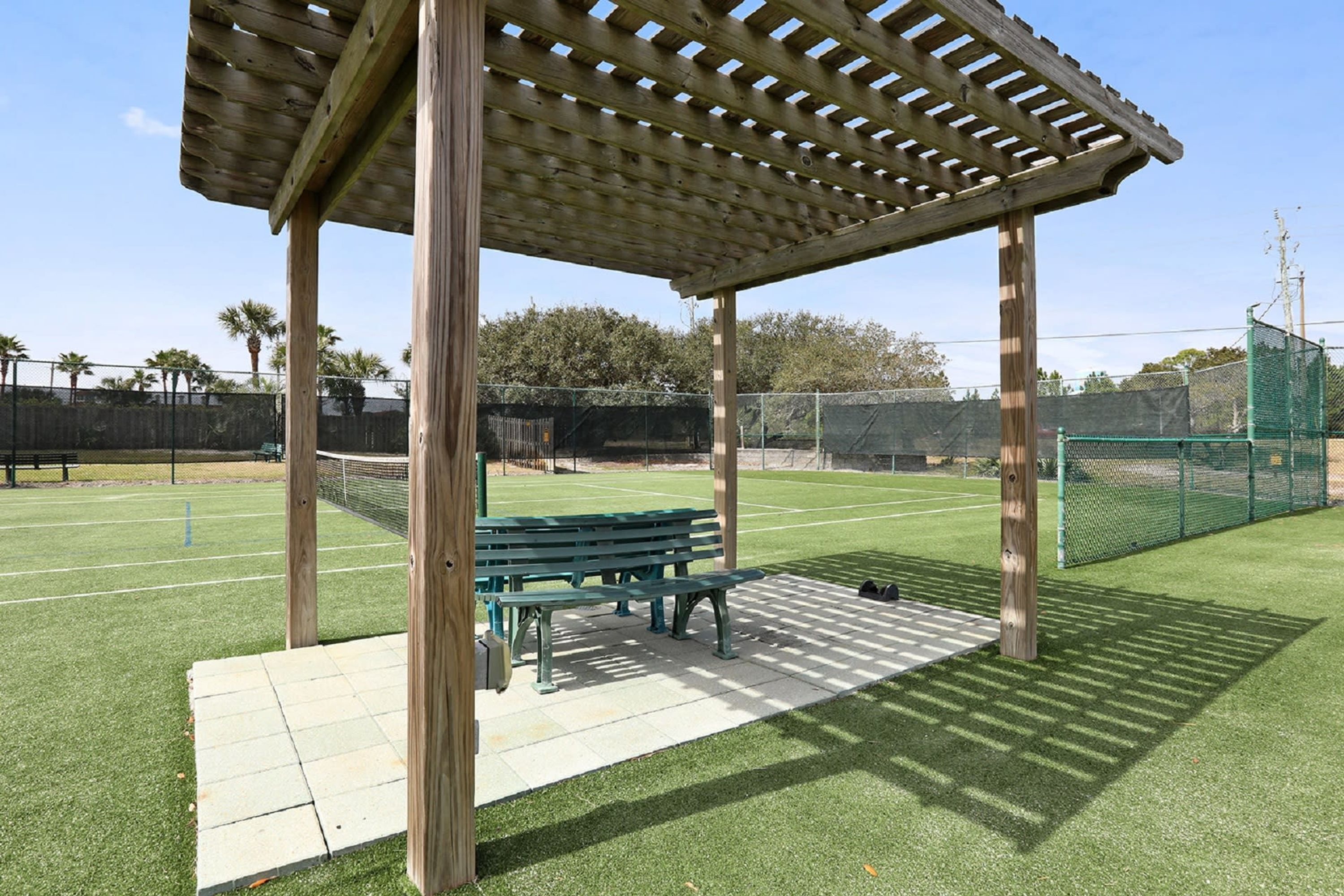 A wooden pergola with benches on a grassy field, surrounded by a fence.