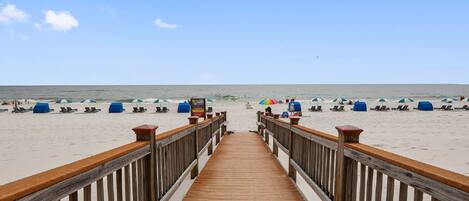 A wooden pier leading to a beach with blue umbrellas and lounge chairs.