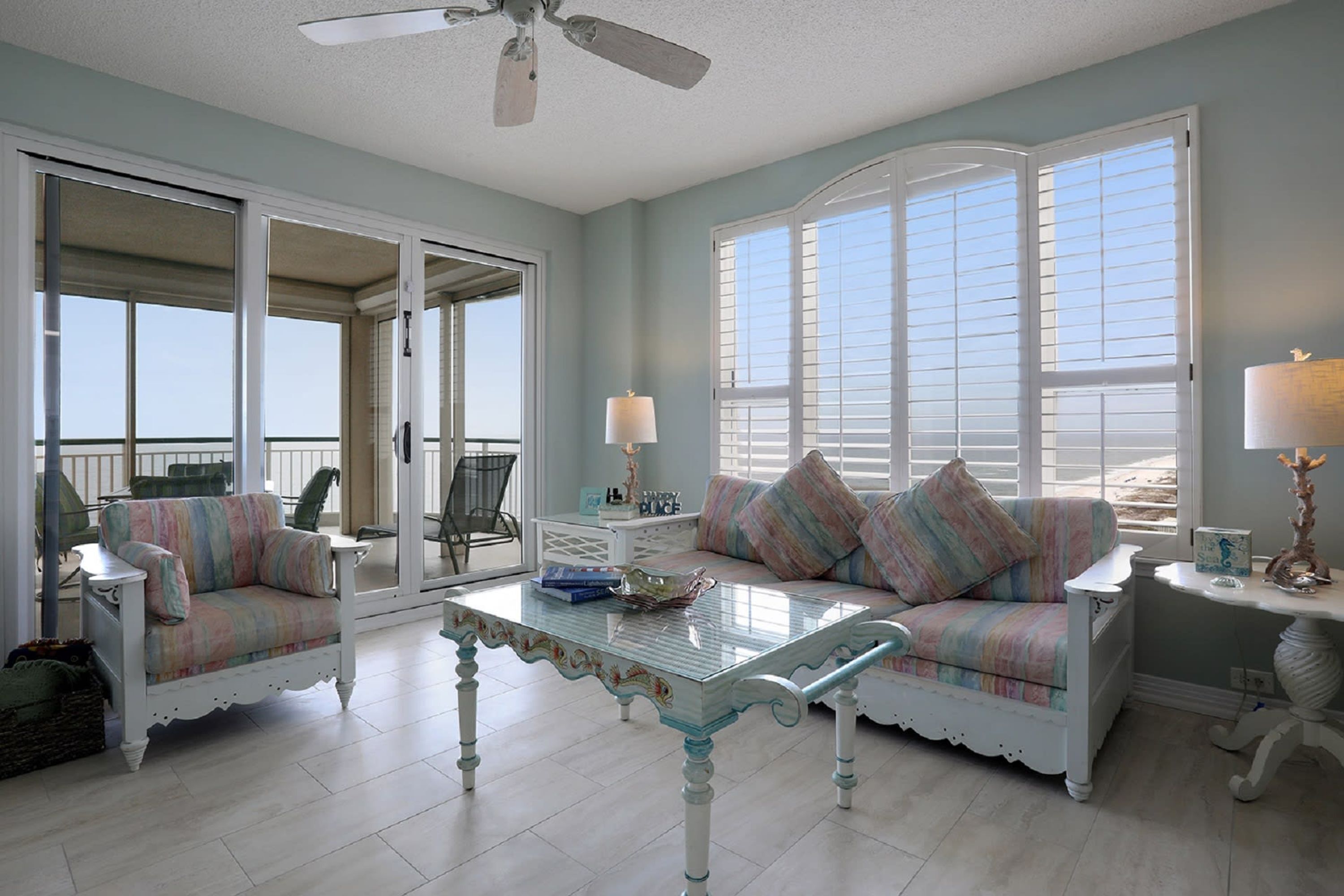 A spacious room with a large sliding glass door leading to a balcony, a white sofa with striped cushions, a glass coffee table, and a white side table with a lamp.