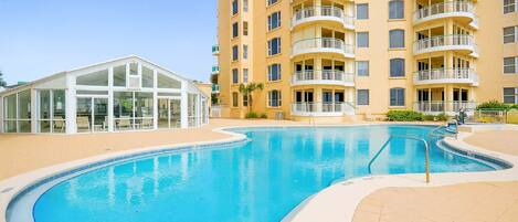 A swimming pool with a curved design, surrounded by a beige tiled area and a multi-story building with balconies.