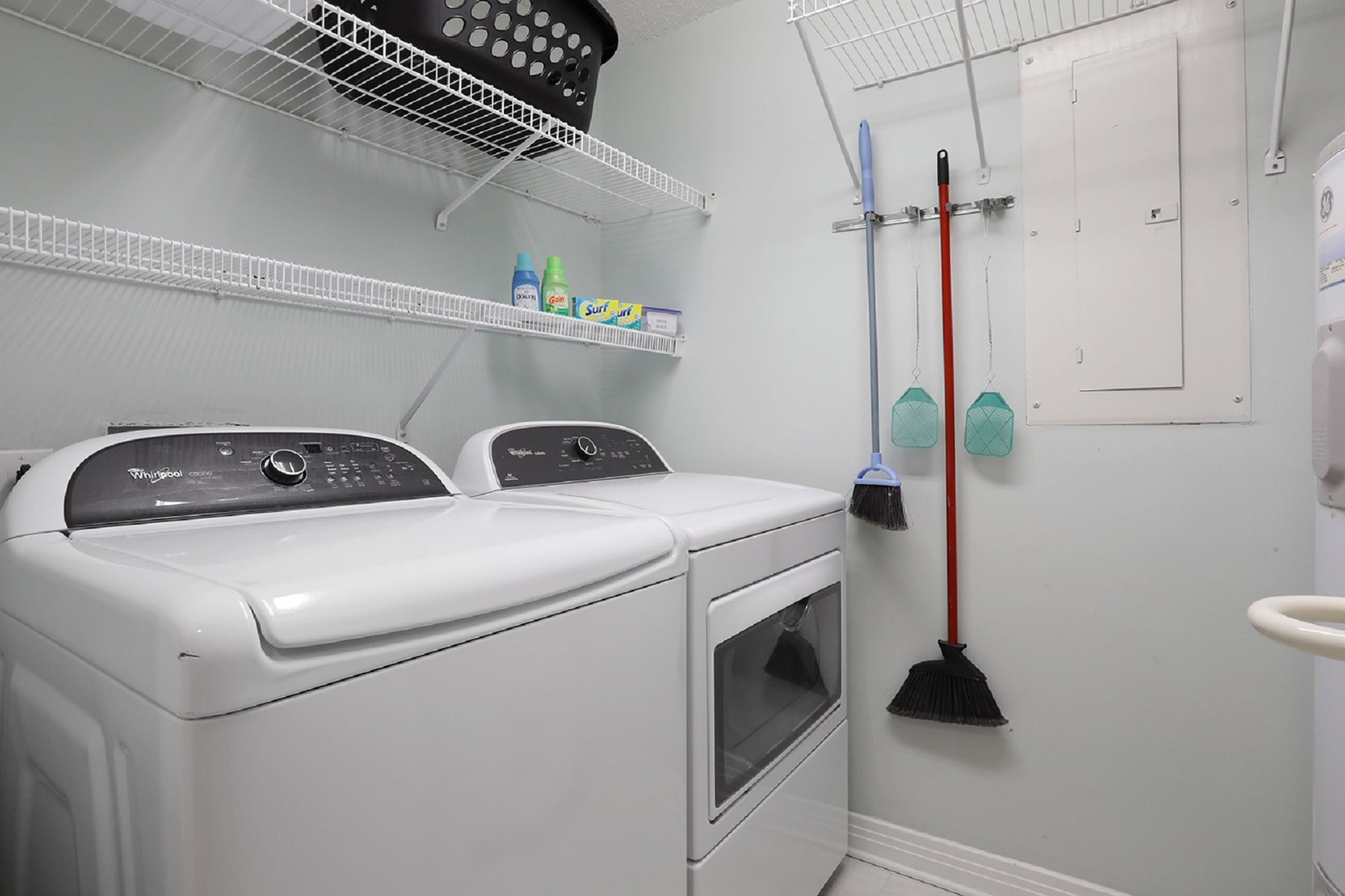 A laundry room with two front-loading washing machines, a shelf with cleaning supplies, and a broom hanging on the wall.