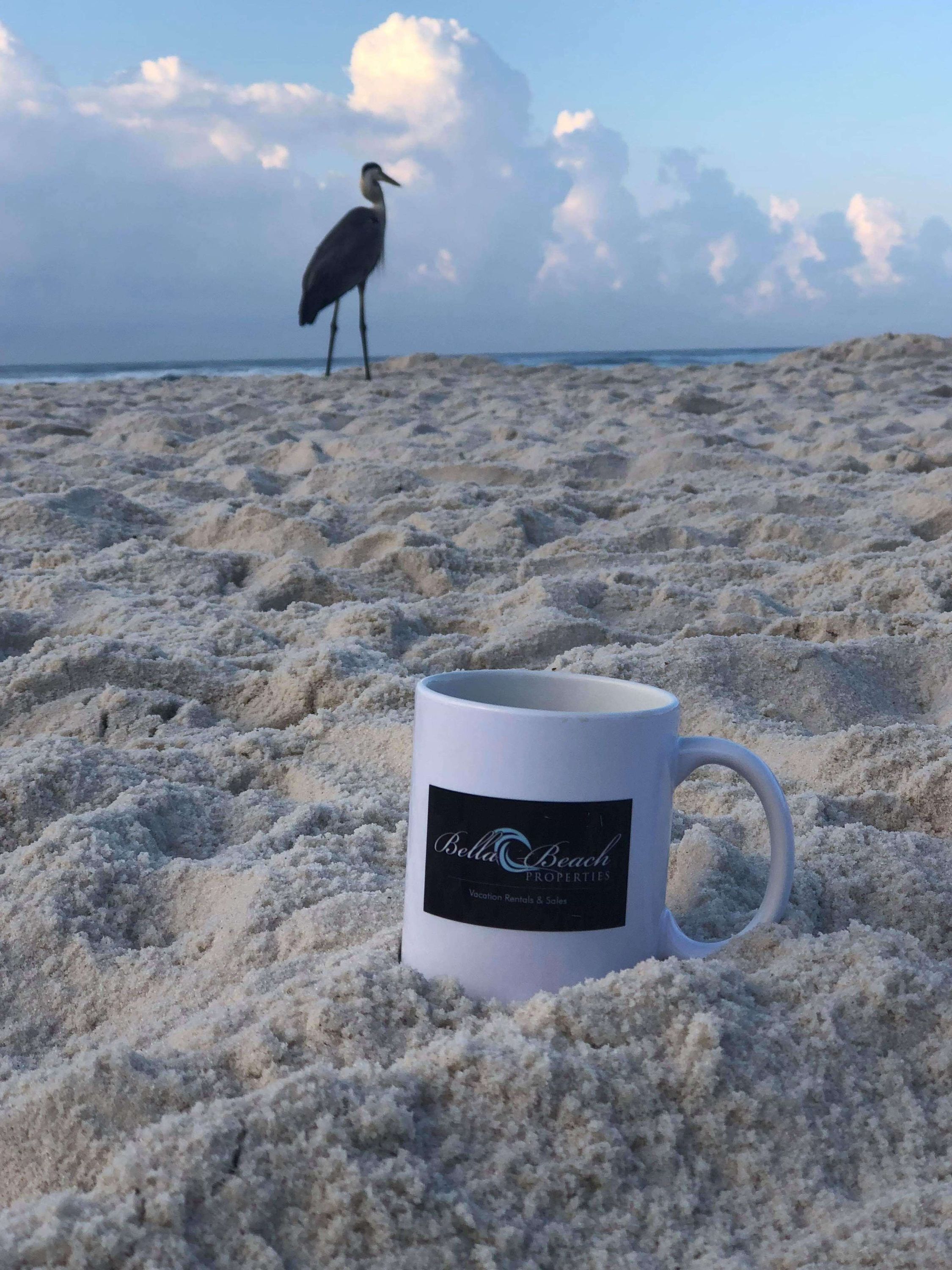 A white mug with a logo on a sandy beach with a bird in the background.