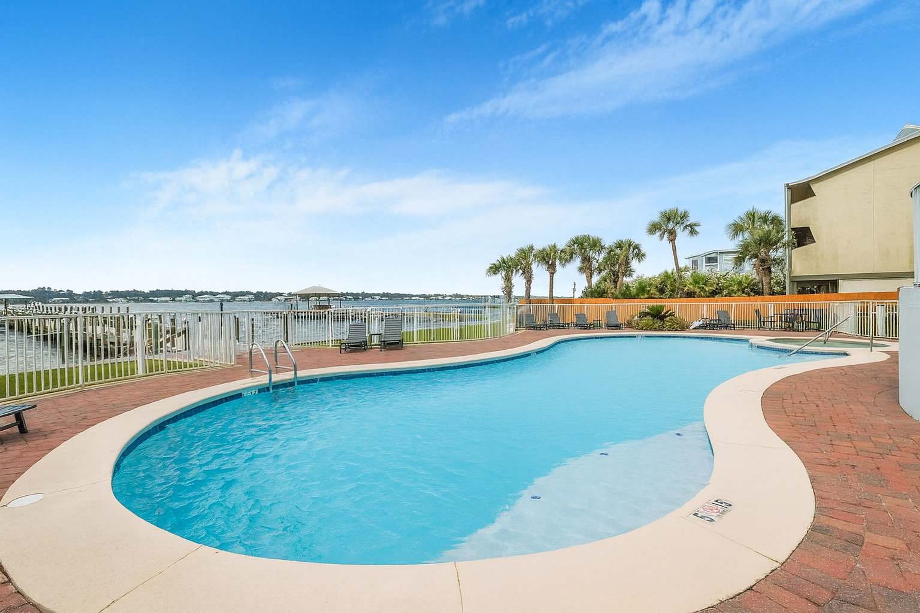 A swimming pool with a curved edge, surrounded by a brick walkway, palm trees, and a view of the ocean.