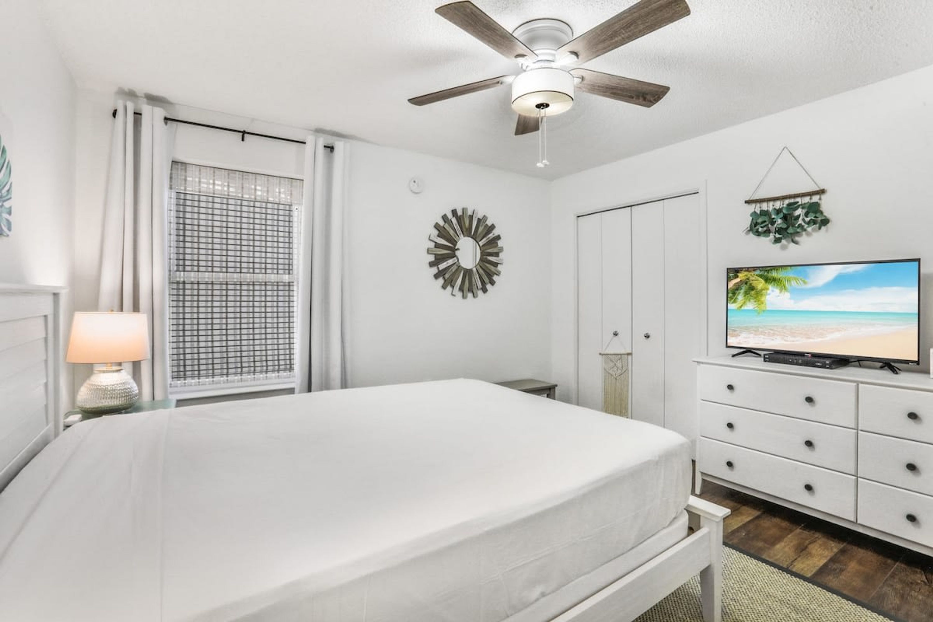 A bedroom with a bed, a ceiling fan, a TV, a dresser, and a window with curtains.