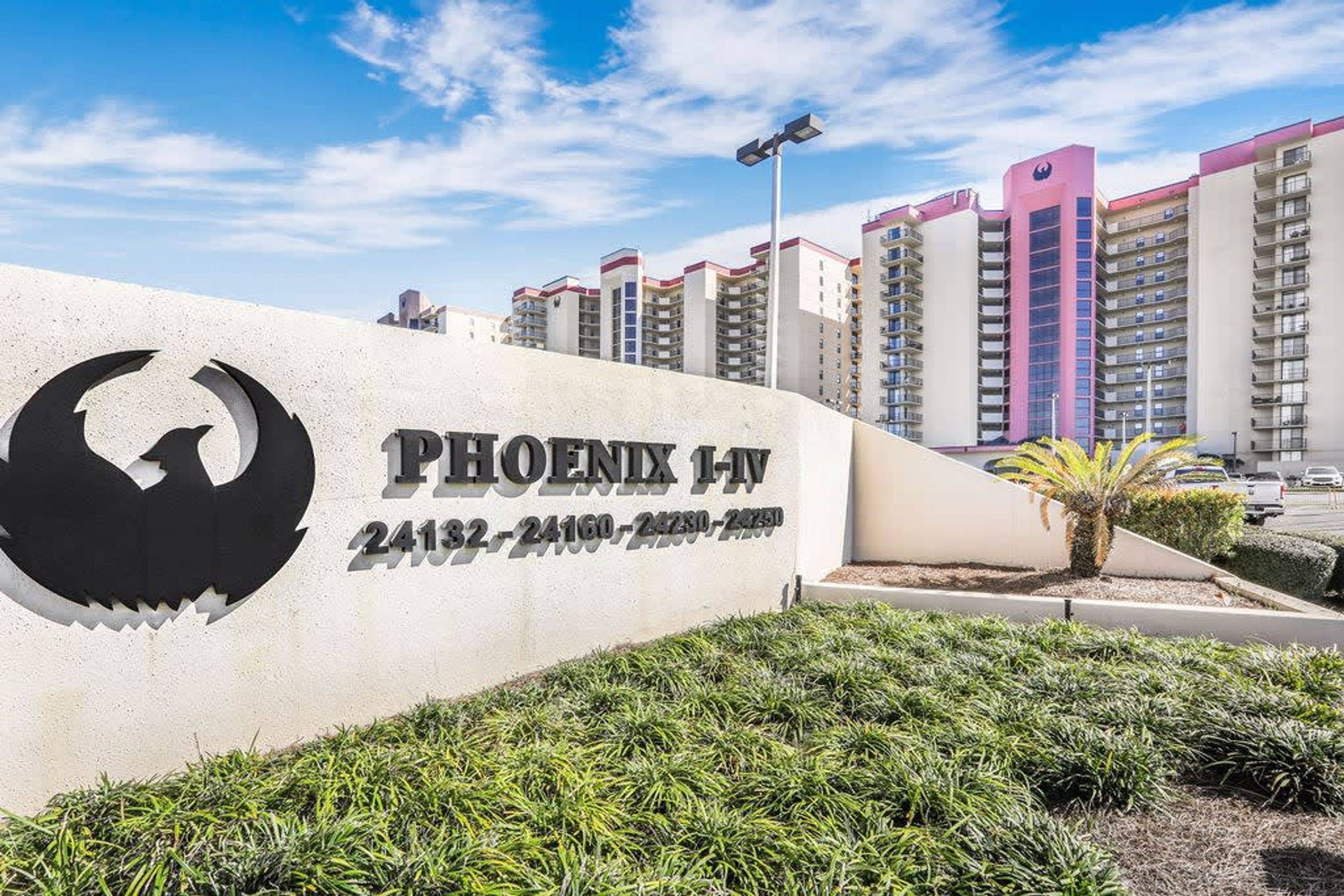 A sign for Phoenix 1-NV with contact numbers, surrounded by greenery and tall buildings in the background.
