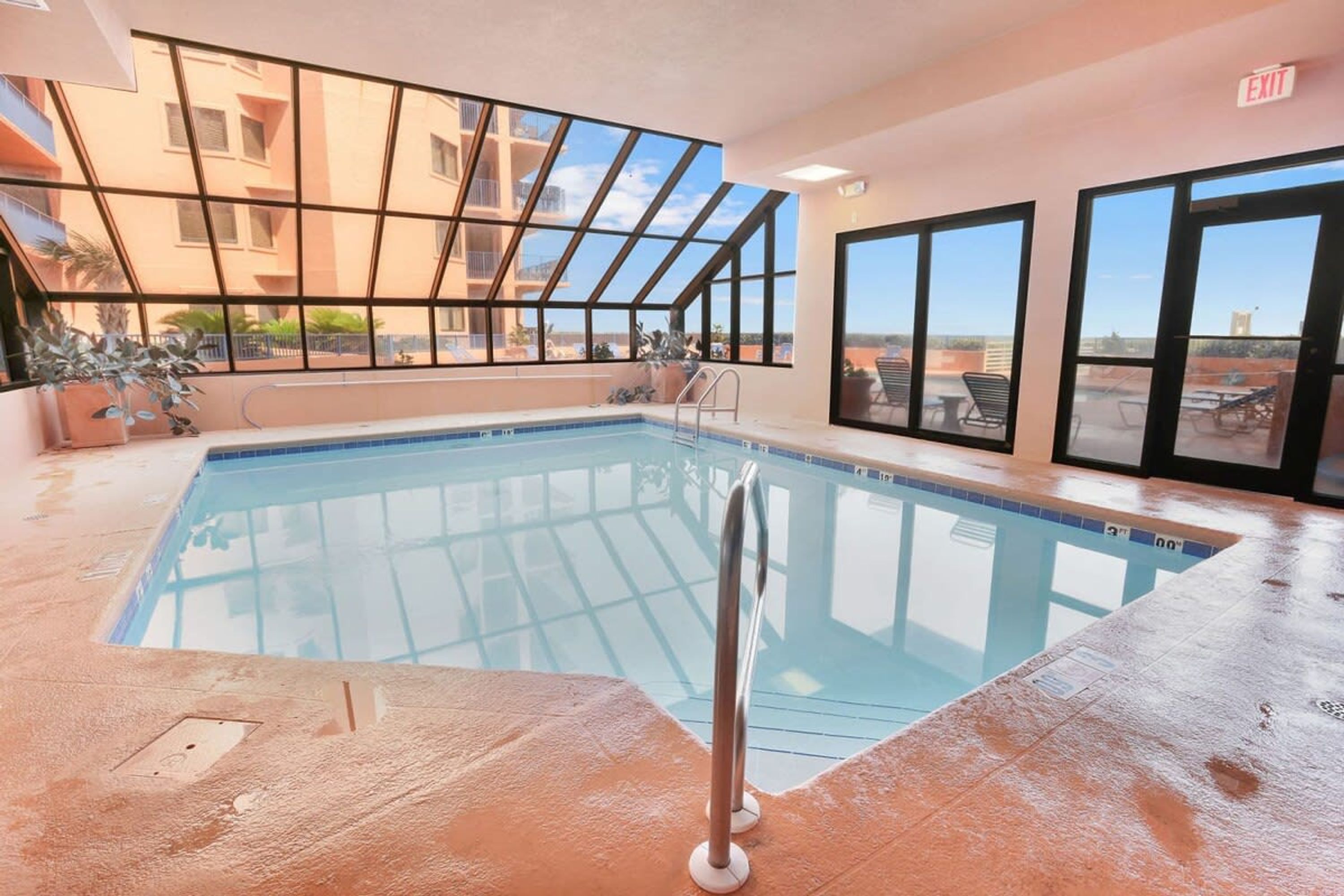 An indoor swimming pool with clear glass walls, a metal ladder, and a view of a building with balconies.