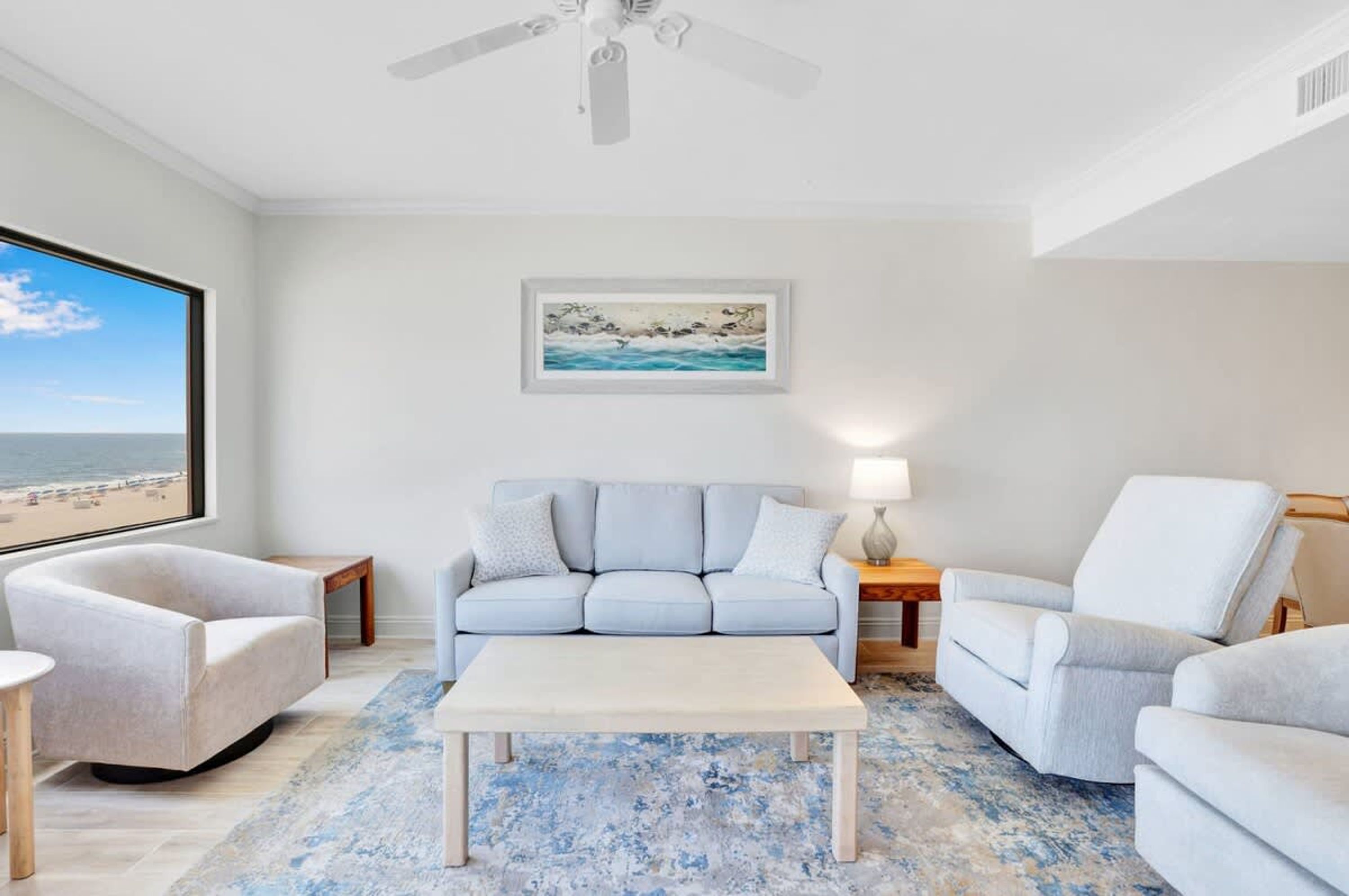 A living room with a sofa, armchairs, a coffee table, a ceiling fan, a painting, and a view of the beach.
