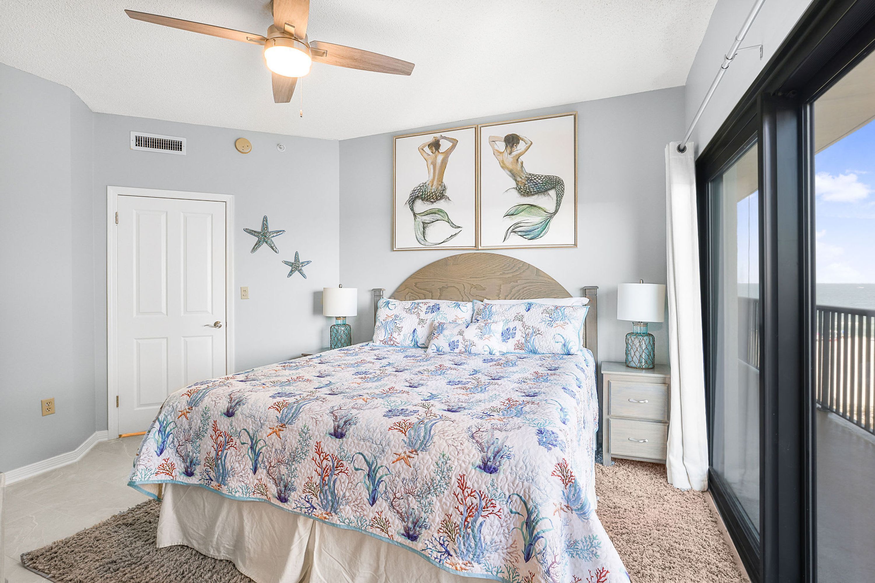 Un dormitorio con cama, ventilador de techo, puerta, dos adornos de estrellas de mar en la pared y una ventana grande.