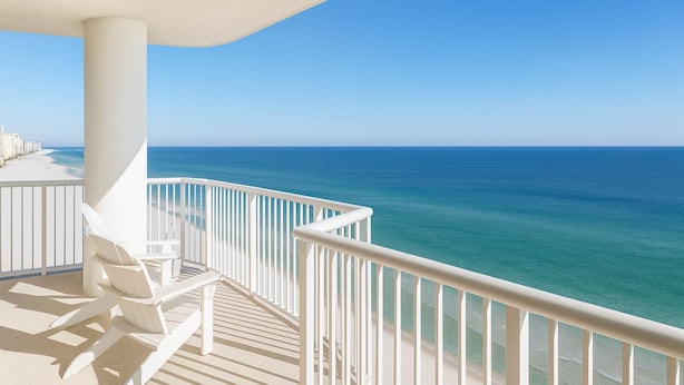 A balcony with white railings, a white umbrella, and a row of white lounge chairs facing a clear blue ocean. - Island Royale- Beachfront Penthouse (Gulf Shores)