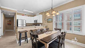 A kitchen with white cabinets, a stainless steel refrigerator, and a microwave. There is a dining area with a wooden table and chairs, and a window with blinds. - Island Royale- Beachfront Penthouse (Gulf Shores)