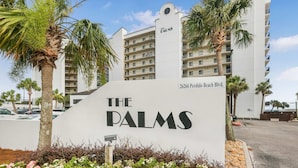 Ein Schild für das Hotel The Palms, mit freier Sicht auf die Außenansicht des Gebäudes und die umliegenden Palmen.