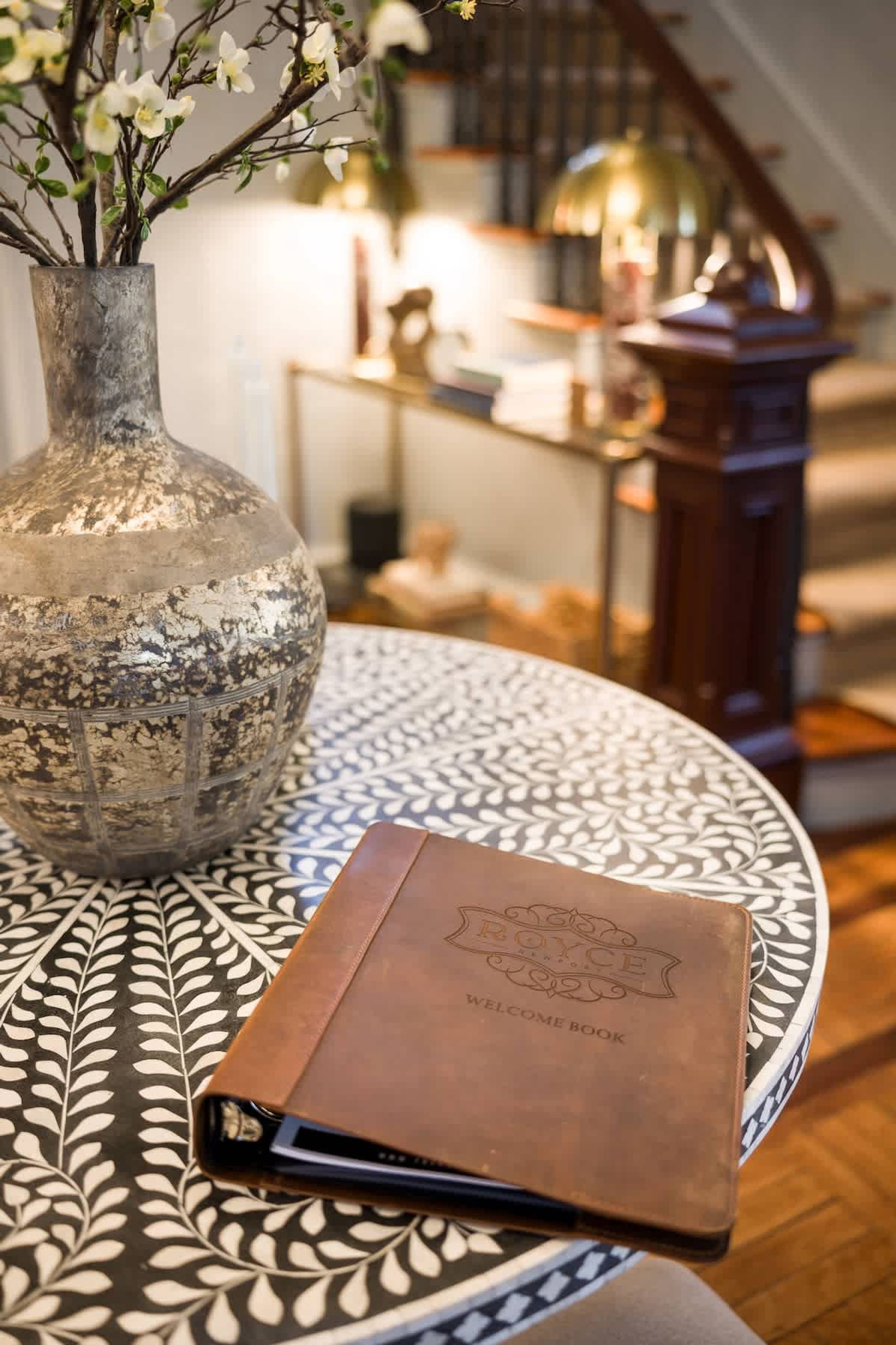 Uma mesa redonda com toalha estampada, um vaso decorativo com flores e um livro de boas-vindas encadernado em couro.