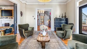 Uma sala de estar ampla com um lustre grande, poltronas estofadas em verde, uma mesa de centro de madeira com um vaso de flores e vista para o interior através de uma porta.