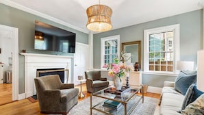 A living room with a fireplace, a television mounted above it, and a chandelier. - Regatta Suite at The Royce Refined Newport Charm (Newport)