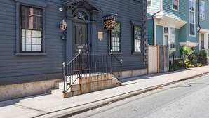 A two-story building with a dark blue exterior, a front porch with a black metal railing, and a sign that reads "ROYCE". - Regatta Suite at The Royce Refined Newport Charm (Newport)