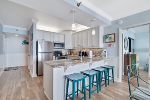 A modern kitchen with white cabinets, a granite island, teal bar stools, and pendant lighting. - Shoalwater 705 Beachfront Condo Pool Hot Tub More (Orange Beach)