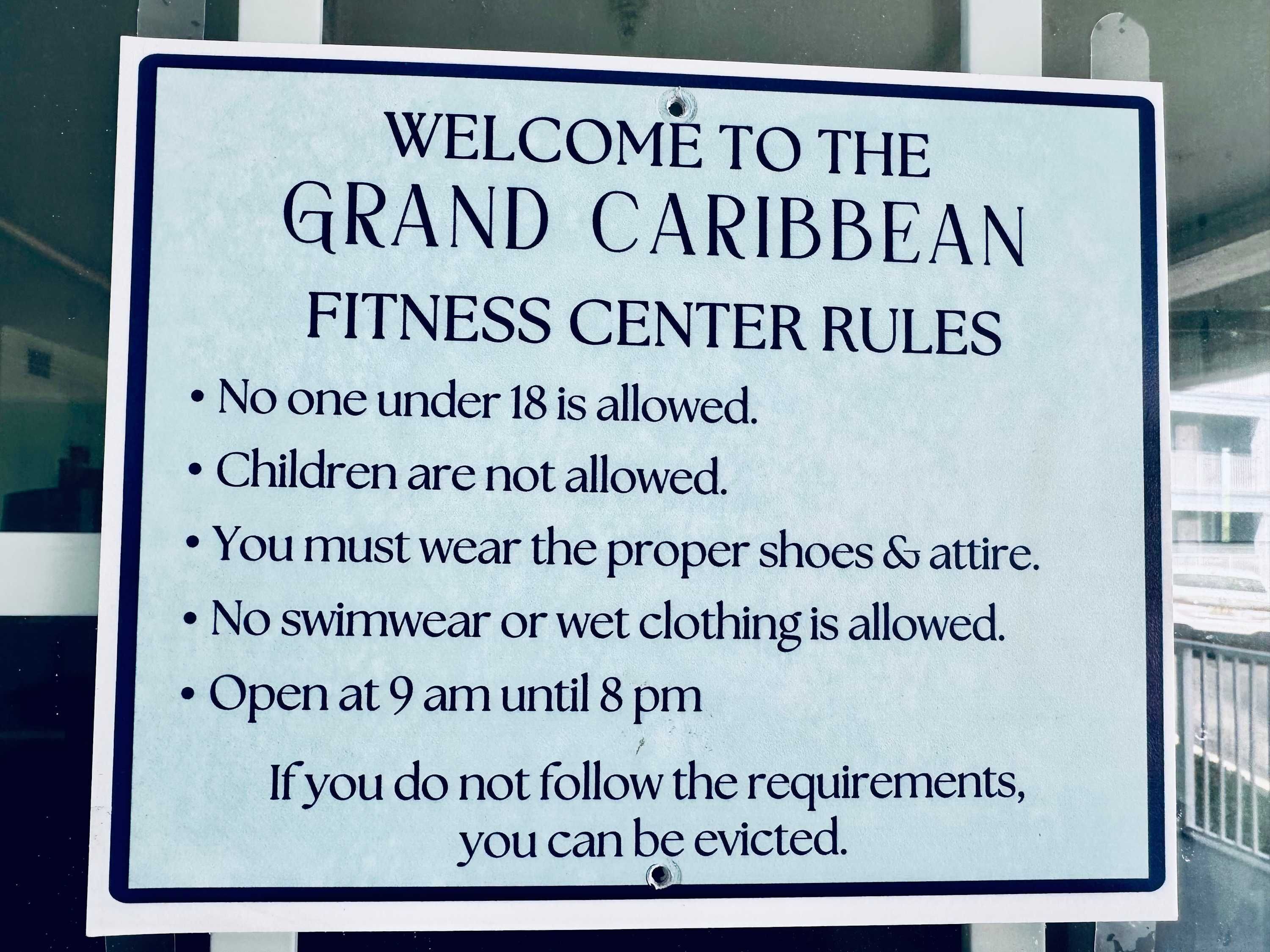 Letrero que enumera las normas del Grand Caribbean Fitness Center, incluyendo restricciones de edad, requisitos de vestimenta y horario de atención.
