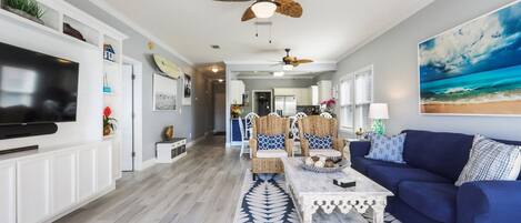 A spacious living room with a large flat-screen TV, a ceiling fan, and a beach-themed painting.
