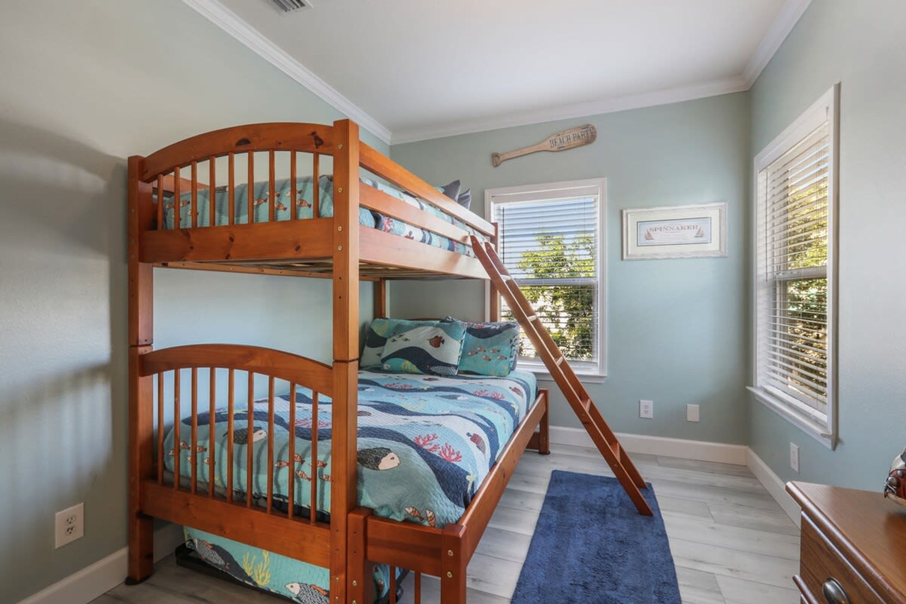 Un lit superposé avec une literie aux motifs bleus et blancs évoquant l’océan, une échelle en bois et un tapis bleu sur un plancher de couleur claire.