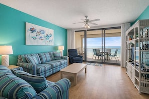 A living room with a teal wall, striped sofa, a coffee table, a ceiling fan, and a balcony with a view of the ocean. - Windward Pointe 903- Beachfront Indoor Outdoor Pool (Orange Beach)