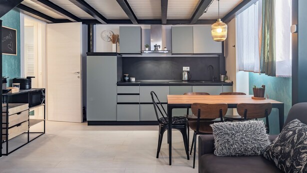 A modern kitchen with grey cabinets, a wooden dining table, and black chairs. There is a black-framed sofa with patterned cushions in the foreground. - Milan Central Station - Green Loft (Milano)