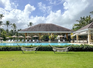 Uma área de piscina com espreguiçadeiras e guarda-sóis, cercada por palmeiras e um pavilhão coberto.