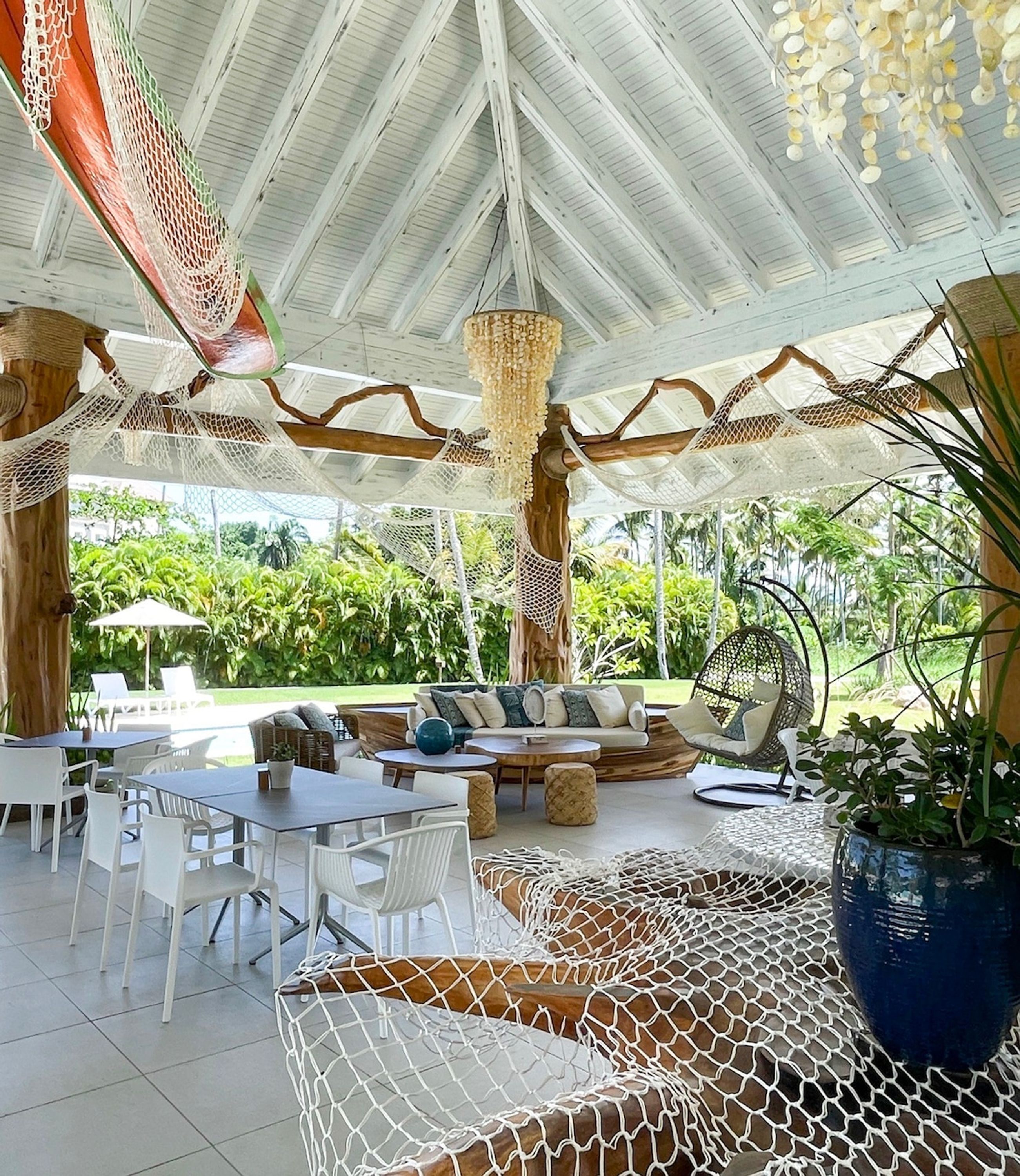 Une terrasse extérieure dotée d’un espace salon couvert, de tables et de chaises, le tout entouré d’une végétation luxuriante.