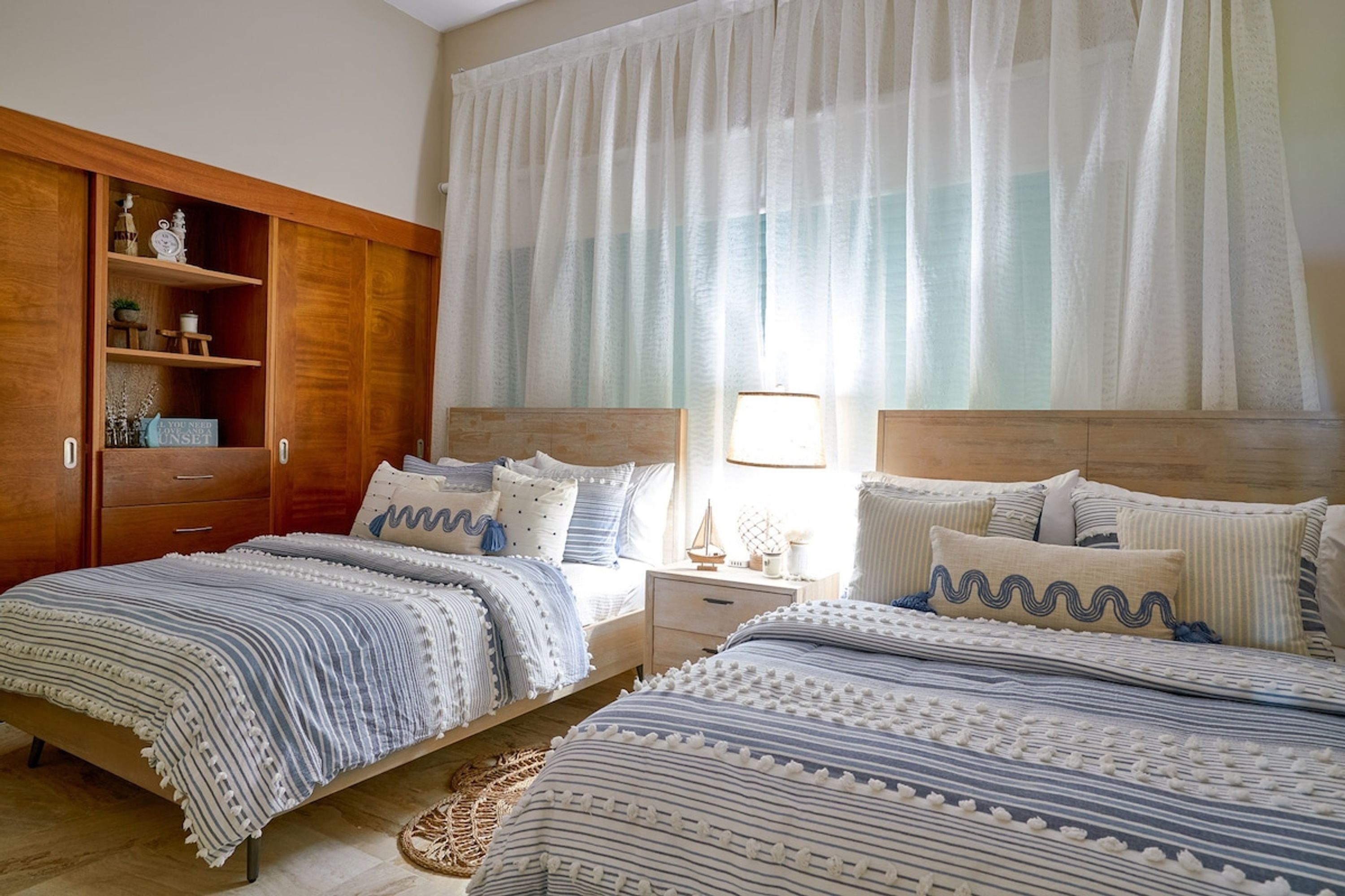 A bedroom with two beds, wooden wardrobe, and a window with sheer curtains.