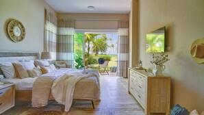 A spacious bedroom with a large bed, a dresser, a vanity, and a television. There is a view of a garden and palm trees outside the window. - The Beach View Playabonita Beachfront Pool (Las Terrenas)