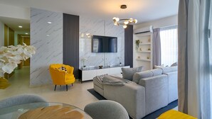 A modern living room with a sofa, a yellow armchair, a glass coffee table, and a TV mounted on the wall. - 2BR The City Center Dtown Pool Gym Nearcecilip (Santo Domingo)